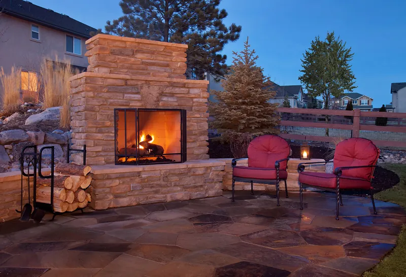 An outdoor masonry fireplace burning on a stone patio at dusk